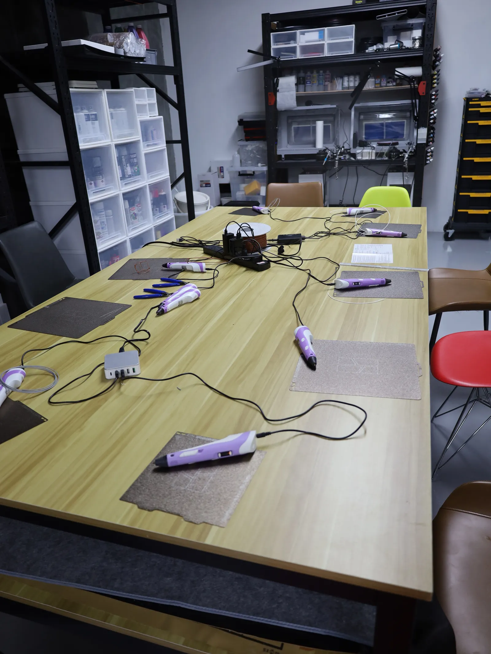 A long studio table set up for the 3D pen station, with a purple 3D pen and a heat-proof mat at each place and the pens wired to a power hub