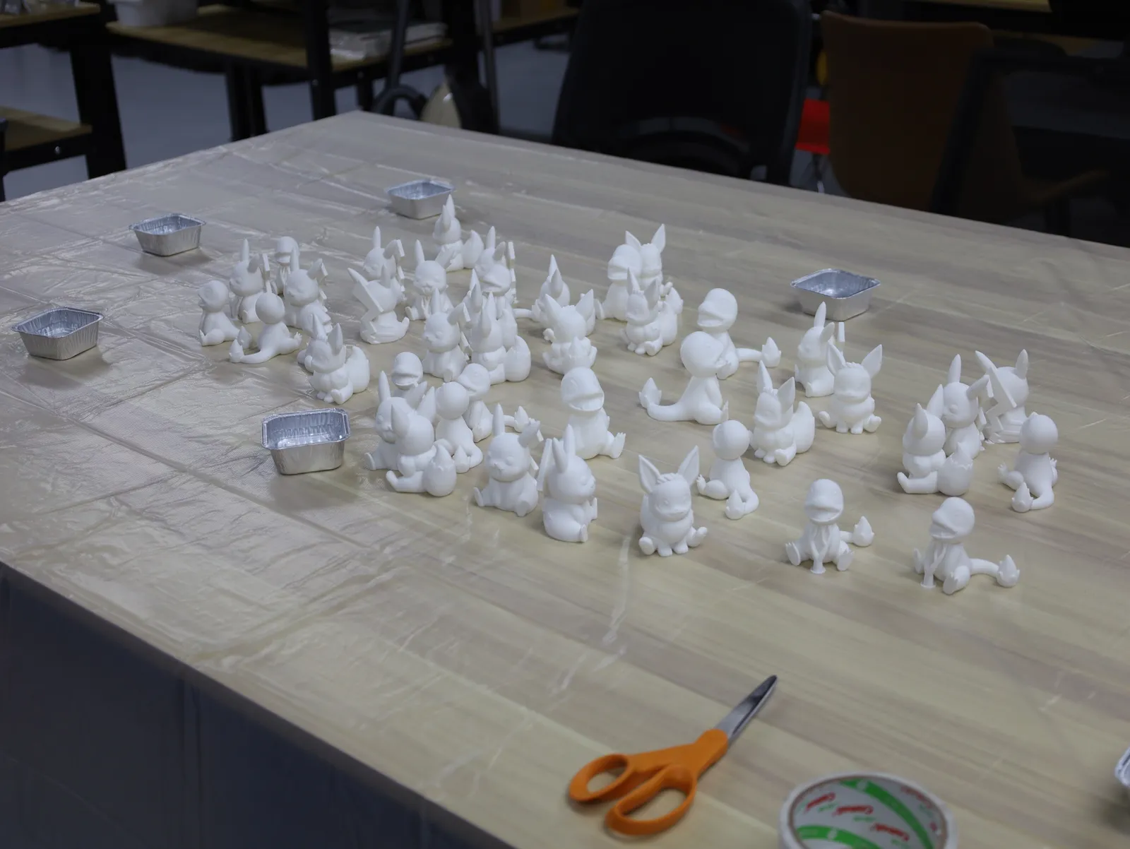 Rows of small white 3D printed figures laid out on a covered table with paint trays, ready for the painting station