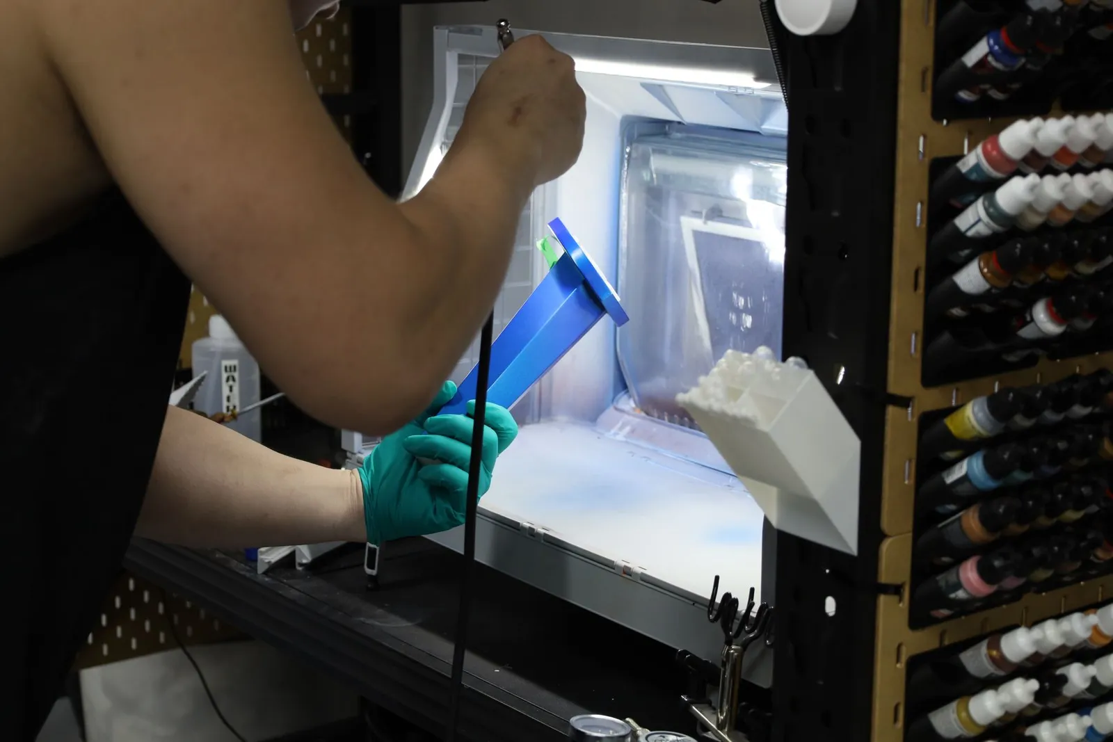 A gloved hand holding a trophy part in the spray booth while an airbrush lays metallic blue over it