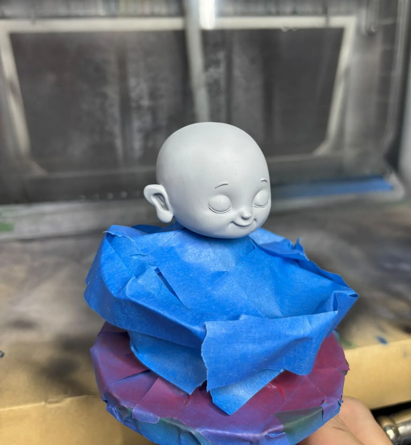 The grey-primed monk figure in the spray booth, its body and base wrapped in blue masking tape so only the head is open to the spray