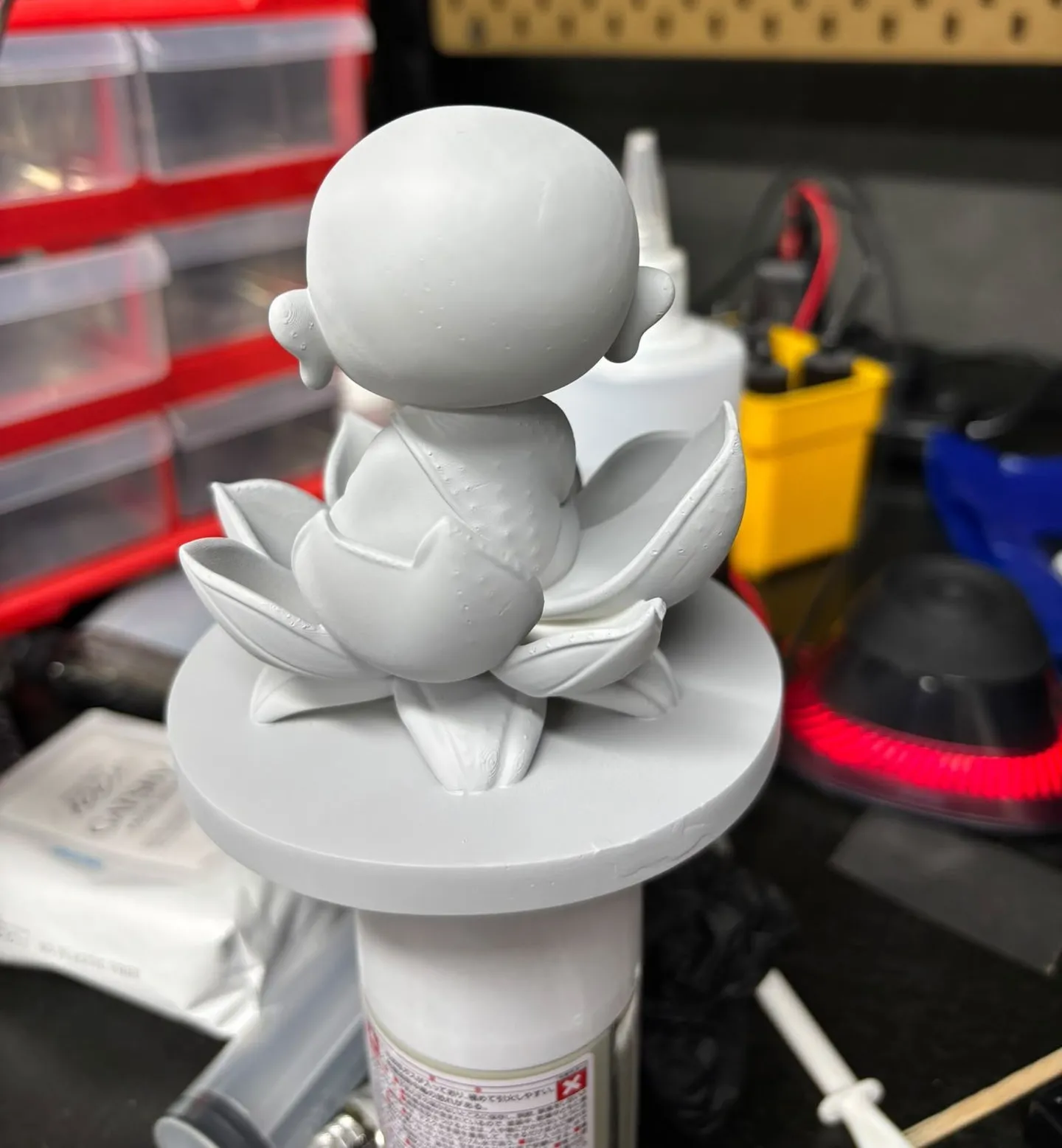 A small monk figure on a lotus base in an even grey primer coat, held up on a spray can on the painting bench
