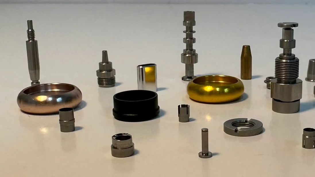 Machined metal parts on a white table: a rose gold ring, a black ring and a gold ring among small steel fittings, pins, a slotted nut and a brass part