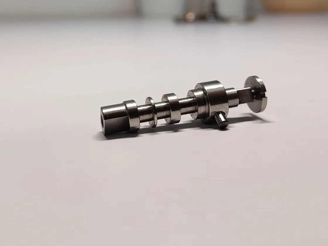 A small machined metal shaft lying on a white table, with stepped collars along its length, a slotted head and a short cross pin