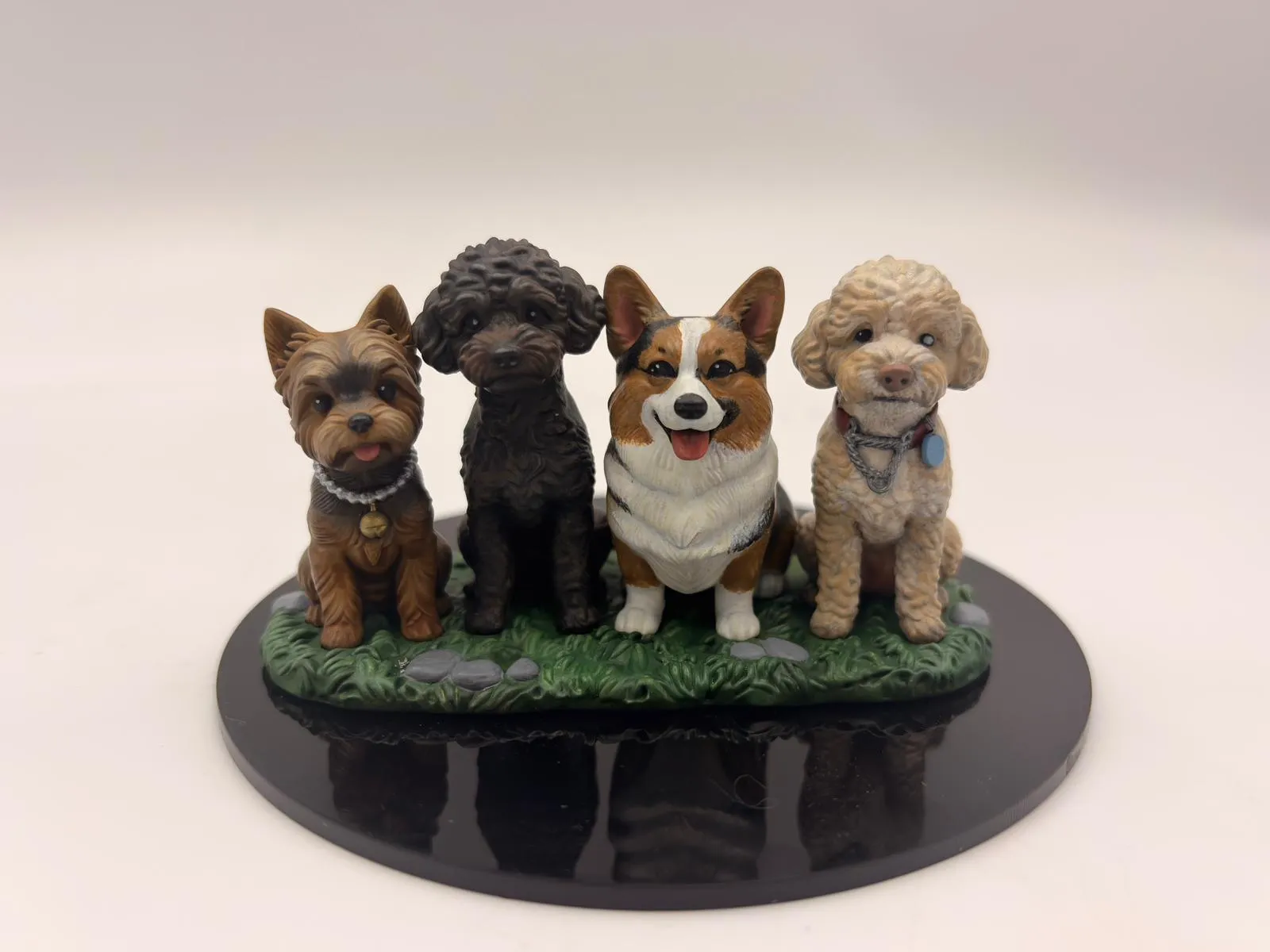 Four hand-painted dog figurines, a terrier, a black poodle, a corgi and a cream poodle, sitting side by side on one grass base