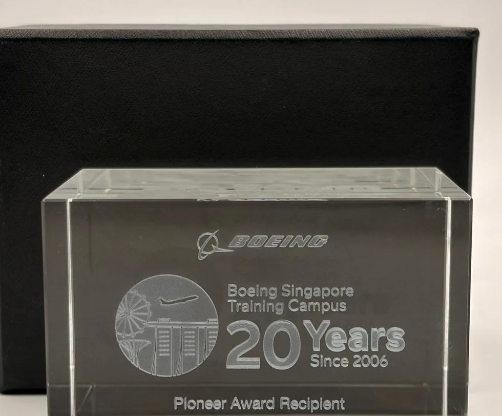 A clear crystal block laser engraved with a company logo, a Singapore skyline roundel and 20 Years Since 2006, standing in front of its black gift box
