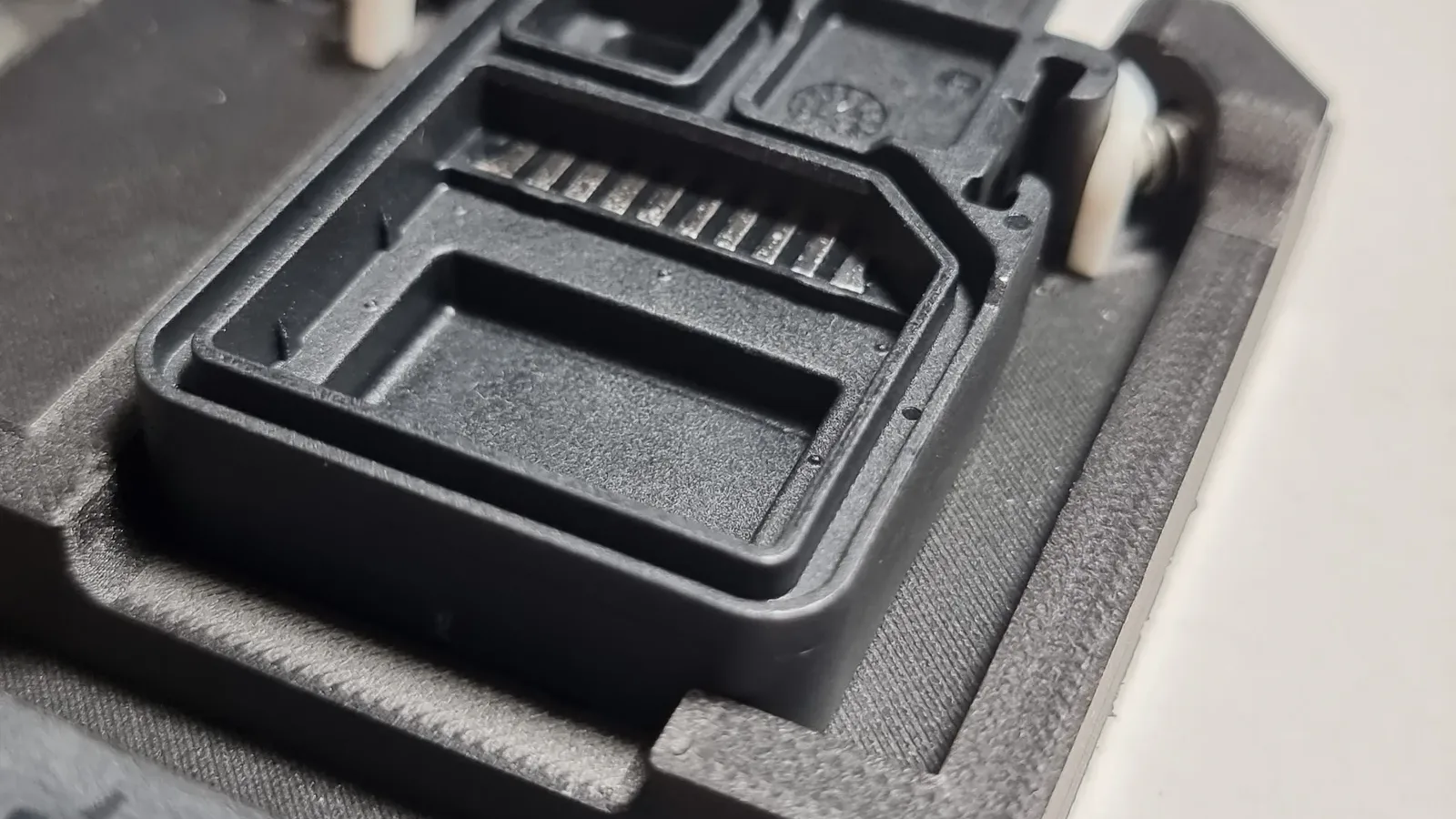 Close-up of an electronic part seated in its pocket in a black 3D printed holding jig, with a white clamp and screw at the edge