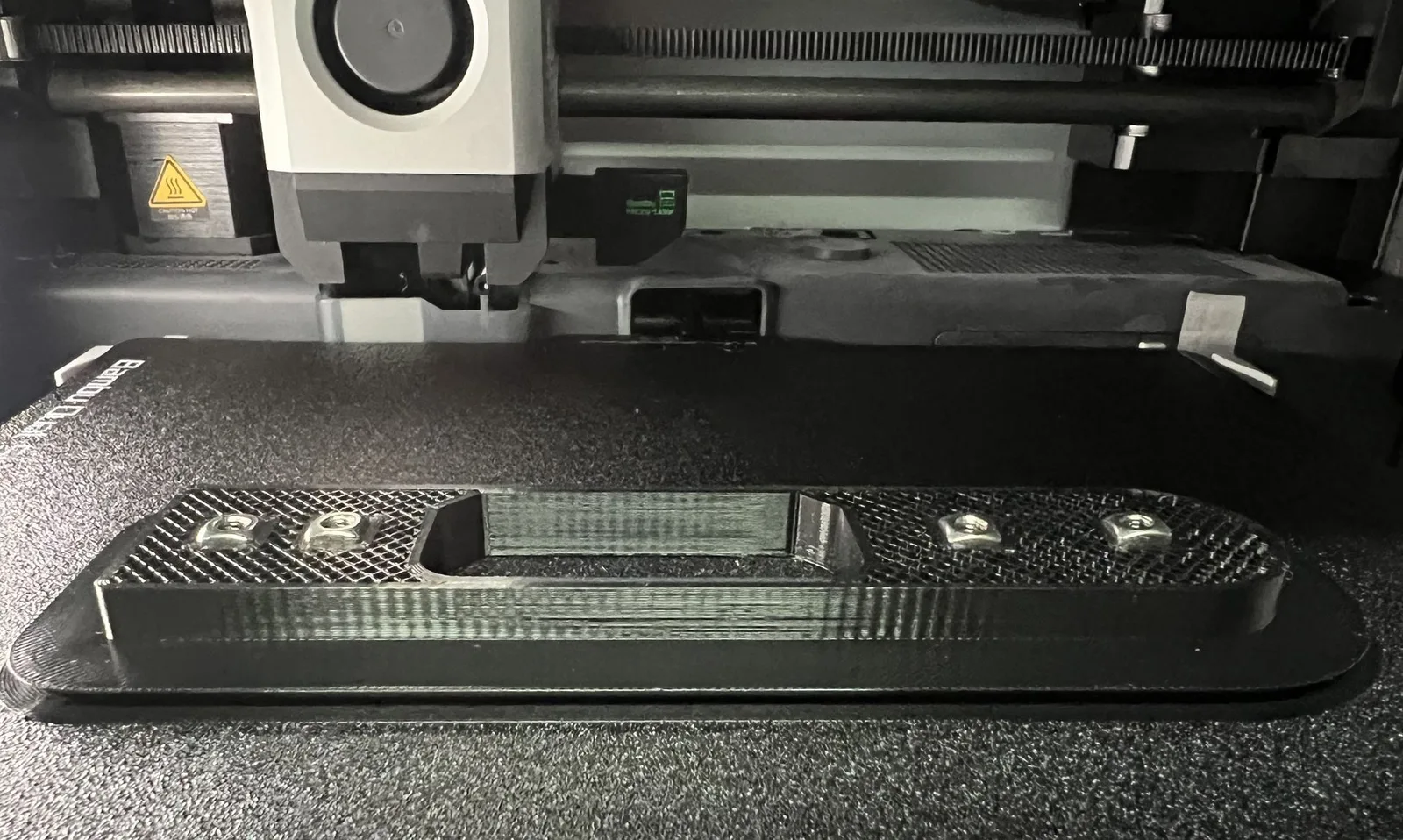Four square nuts sitting in their pockets in a black 3D print, paused mid-print on the printer bed with the infill still open