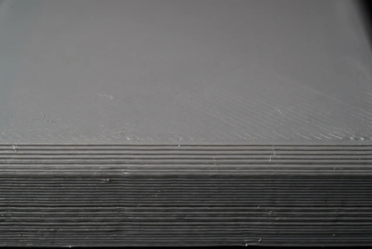 Macro photo of a grey FDM print: raw layered edge with pronounced layer lines below, sanded top surface with only faint traces remaining