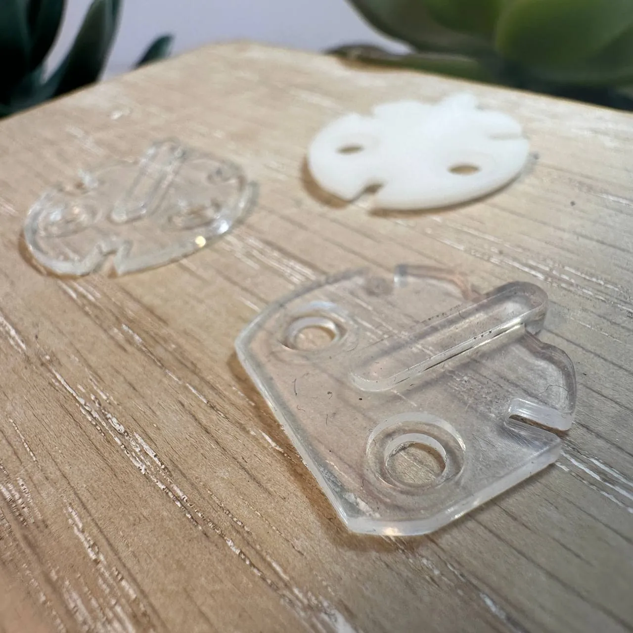 Two clear, flexible 3D printed connectors on a pale wooden board, with the white machined plastic part they were made to replace behind them