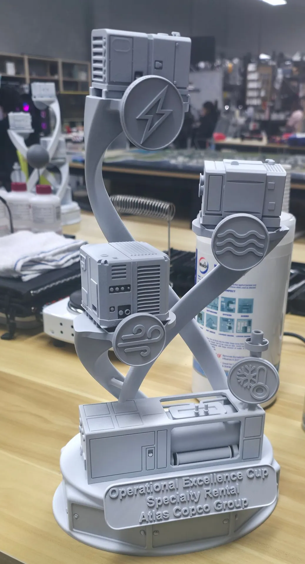 An Operational Excellence Cup trophy in plain grey primer on a workbench, with miniature machines on two twisting arms and a raised plate reading Atlas Copco Group