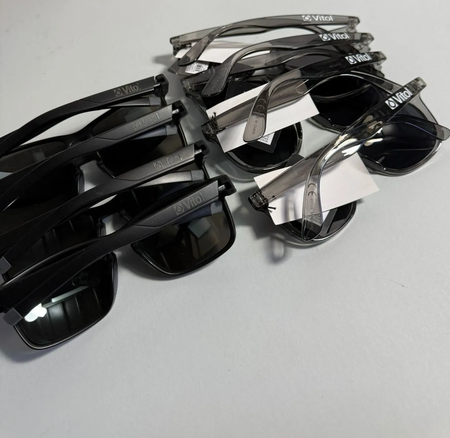 Two sets of branded sunglasses on a white desk: black frames with the Vitol logo engraved in grey on the arms, and clear grey frames with the logo in bright white