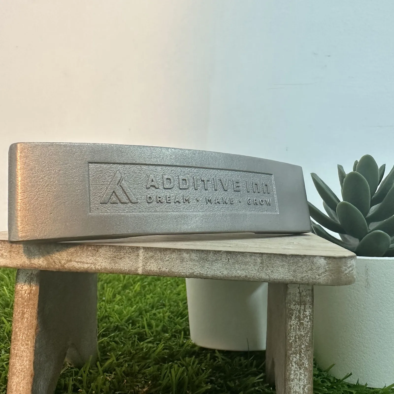 The head of a metal golf putter on a small wooden stand, with the Additive Inn logo and the words Dream, Make, Grow engraved into its face