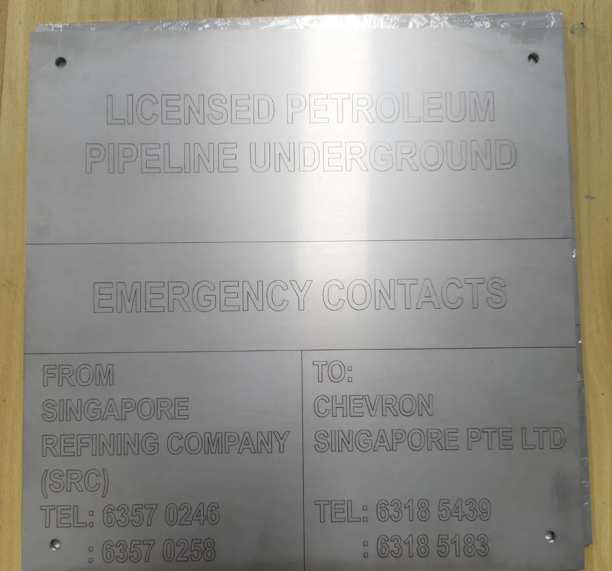 A steel pipeline marker plate laser marked with outlined capital letters reading Licensed Petroleum Pipeline Underground and the emergency contacts below