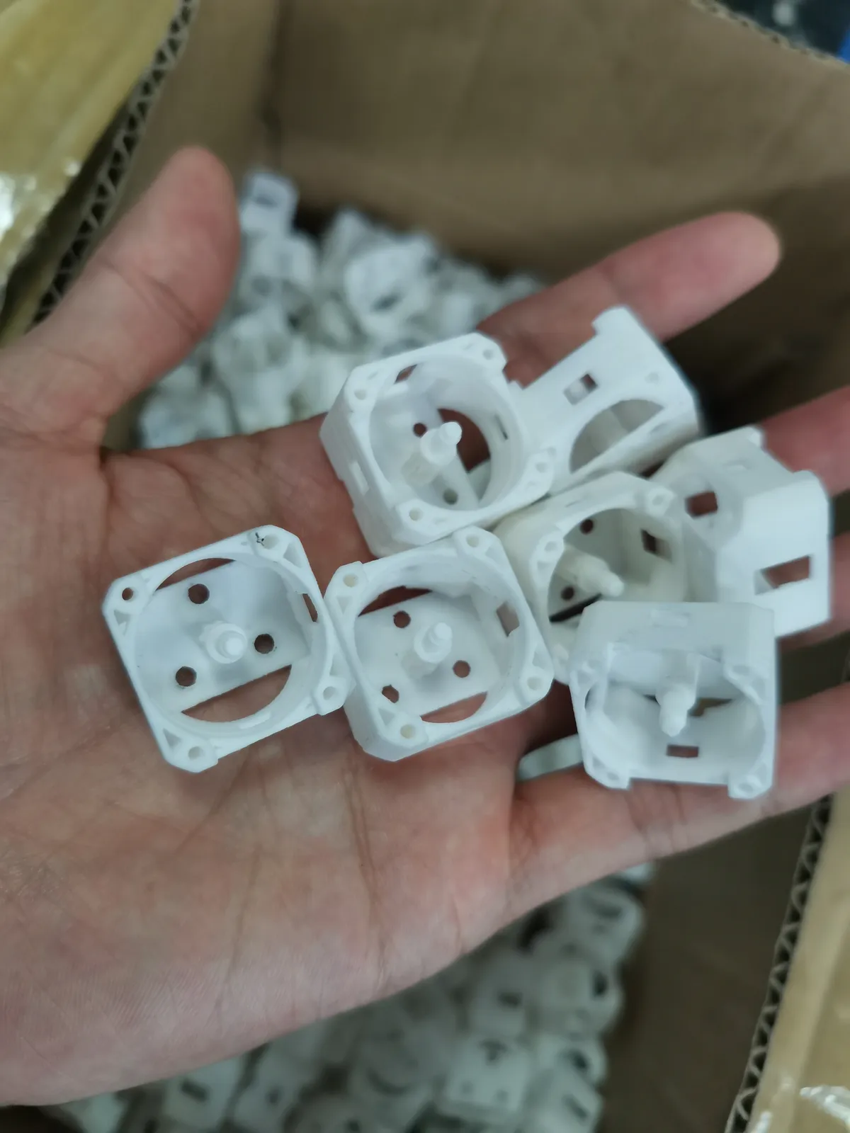 A hand holding a few small white nylon housings printed by SLS, each with a round opening, fixing holes and a central pin, over a box of the same parts