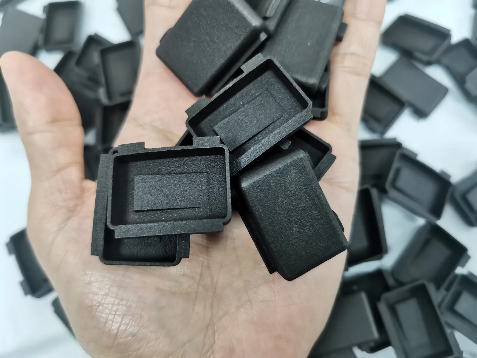 A hand holding black nylon caps printed by SLS, their matte surface showing a fine grain, with more caps scattered on the table