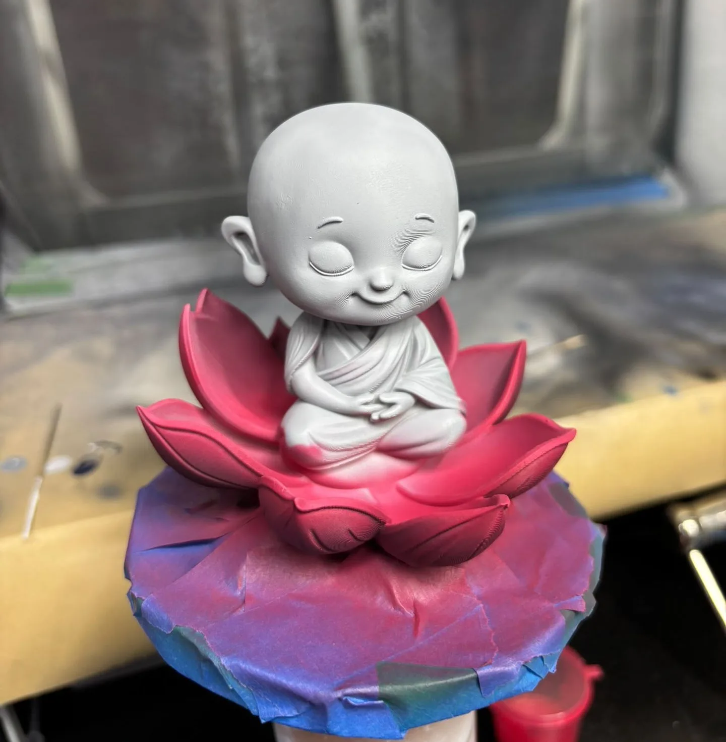 A grey-primed monk figure sitting on a red-sprayed lotus, with red airbrush colour fading up onto the grey robe and tinting the blue masking tape around the base red and purple