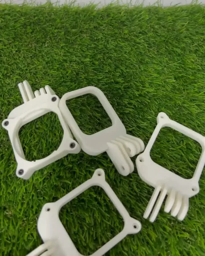 Four white filament-printed camera adapter prototypes on artificial grass, square frames with mounting prongs and screw holes, each a slightly different design