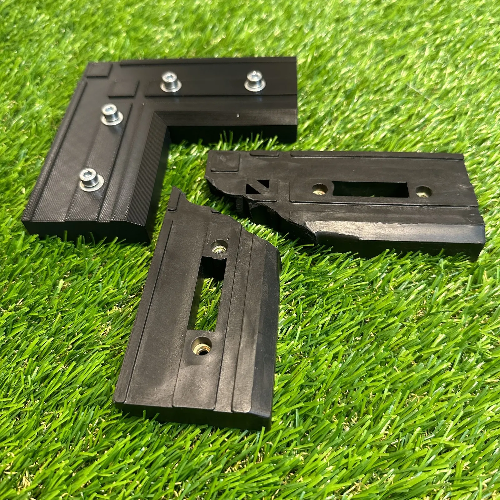 A customer's broken black projector bracket, snapped into two pieces, lying on artificial grass beside the new L-shaped bracket we modelled and 3D printed to replace it, with metal threaded inserts already fitted