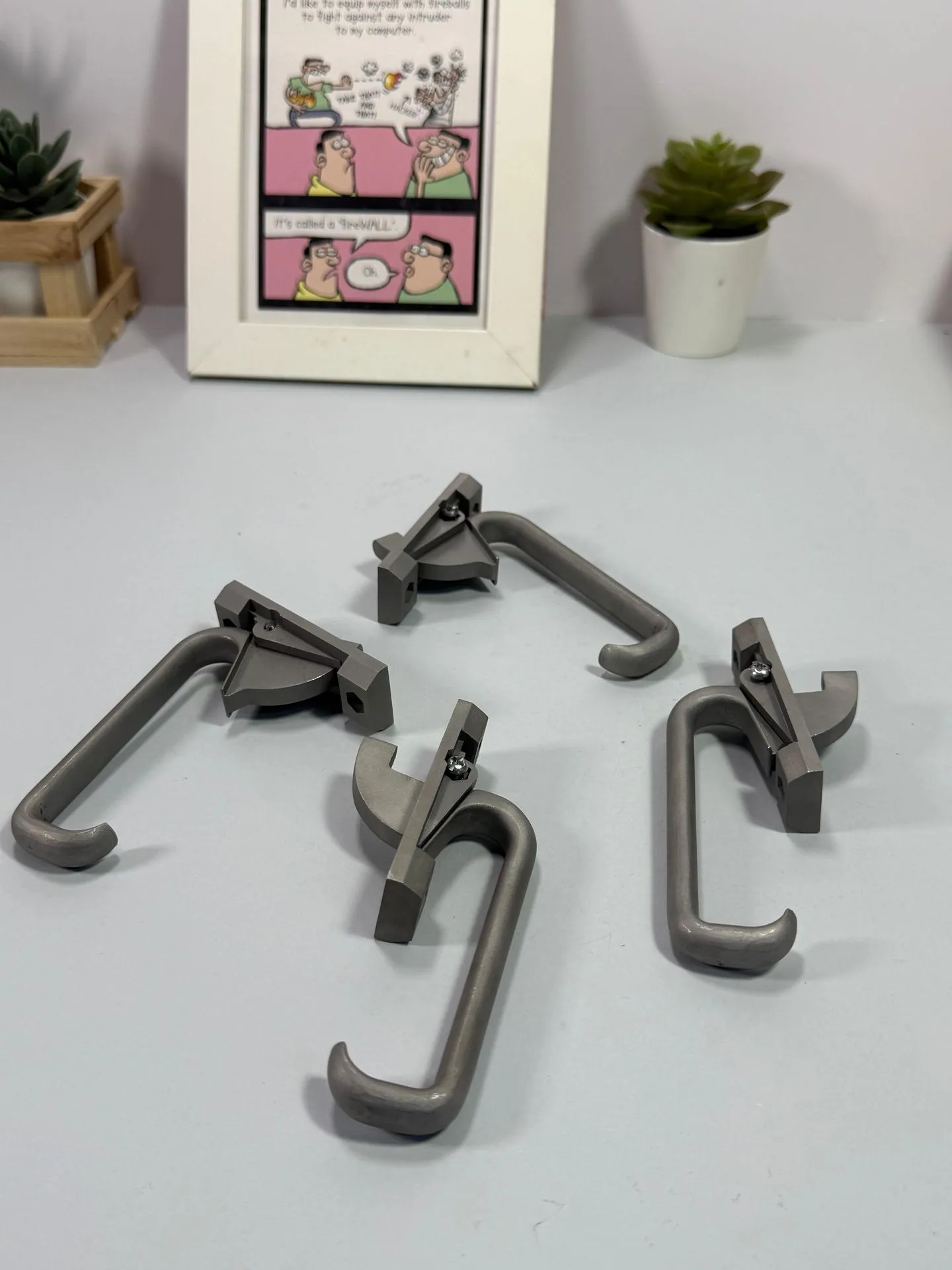 Four grey 3D printed window latches, each with a hooked handle and a small metal pin, laid out on a pale blue table
