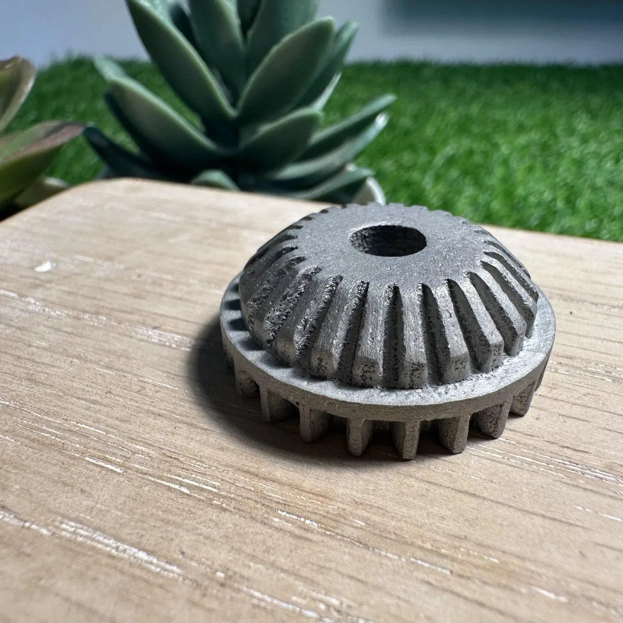A grey metal bevel gear with a centre hole, printed by selective laser melting, sitting on a wooden board, its faces grainy as they came off the machine