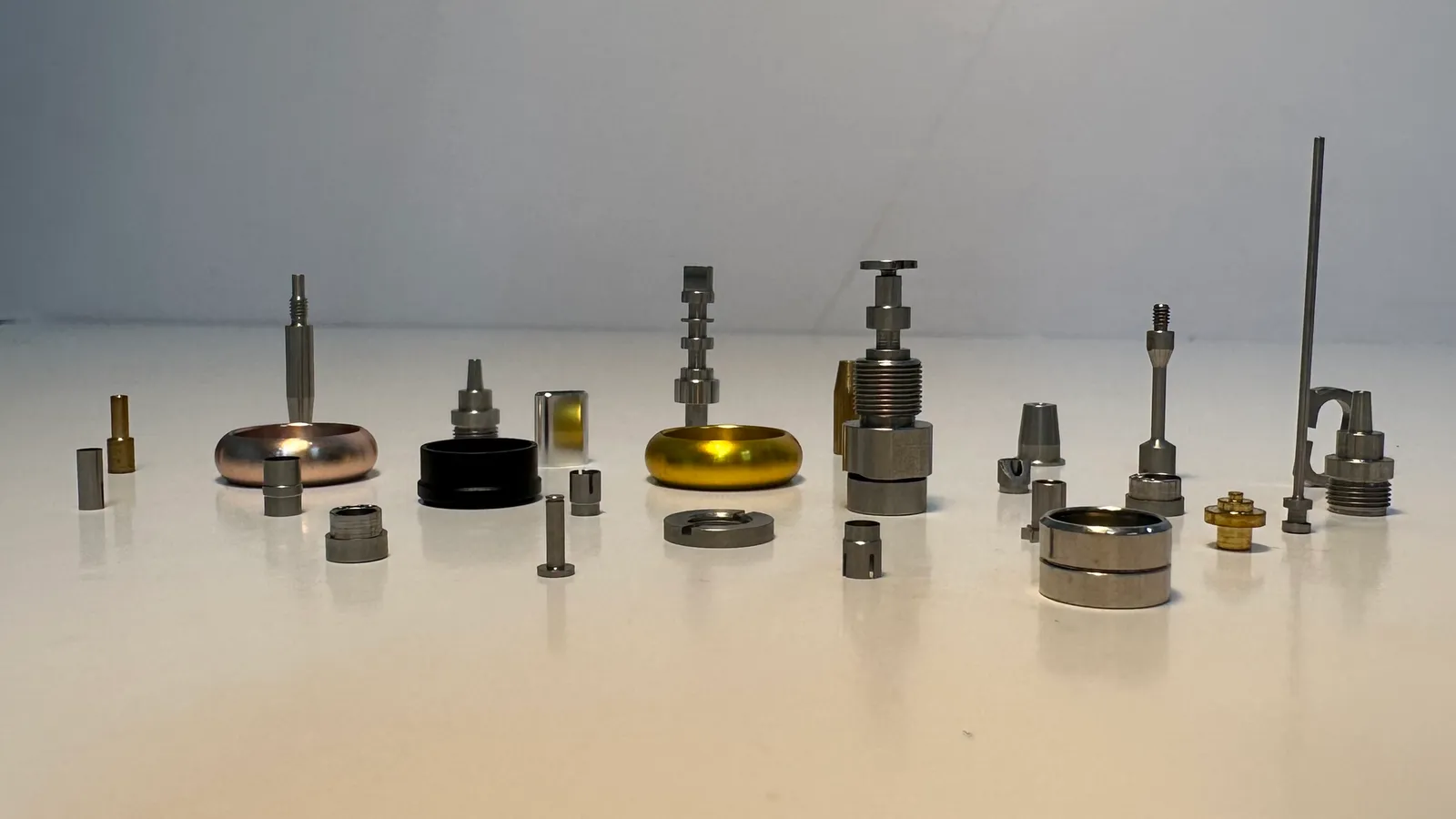 A row of small machined parts on a white table: threaded metal fittings, slim pins, small gold-coloured parts, and rings in rose gold, black and gold