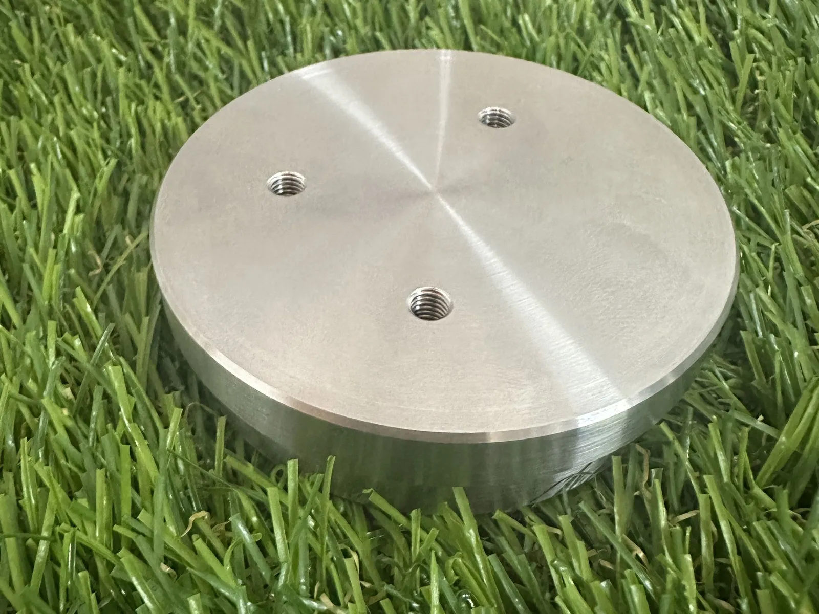 A round machined metal disc on artificial grass, its top face showing the fine circular tool marks of turning, with three threaded screw holes