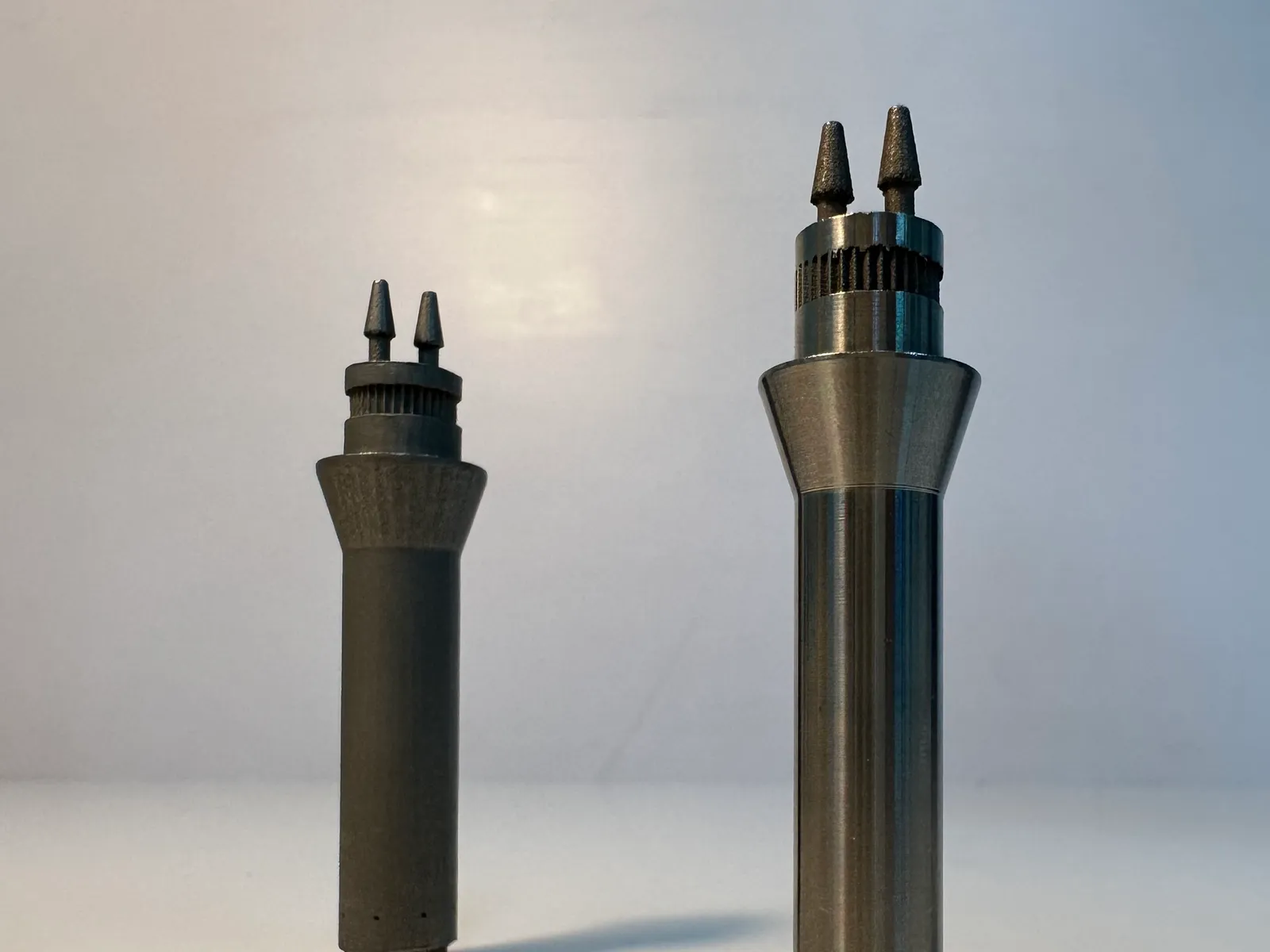 Two metal nozzles side by side: the left one dull grey and slightly rough all over, the right one with a bright, smooth machined body below rough barbed outlets and a ribbed band