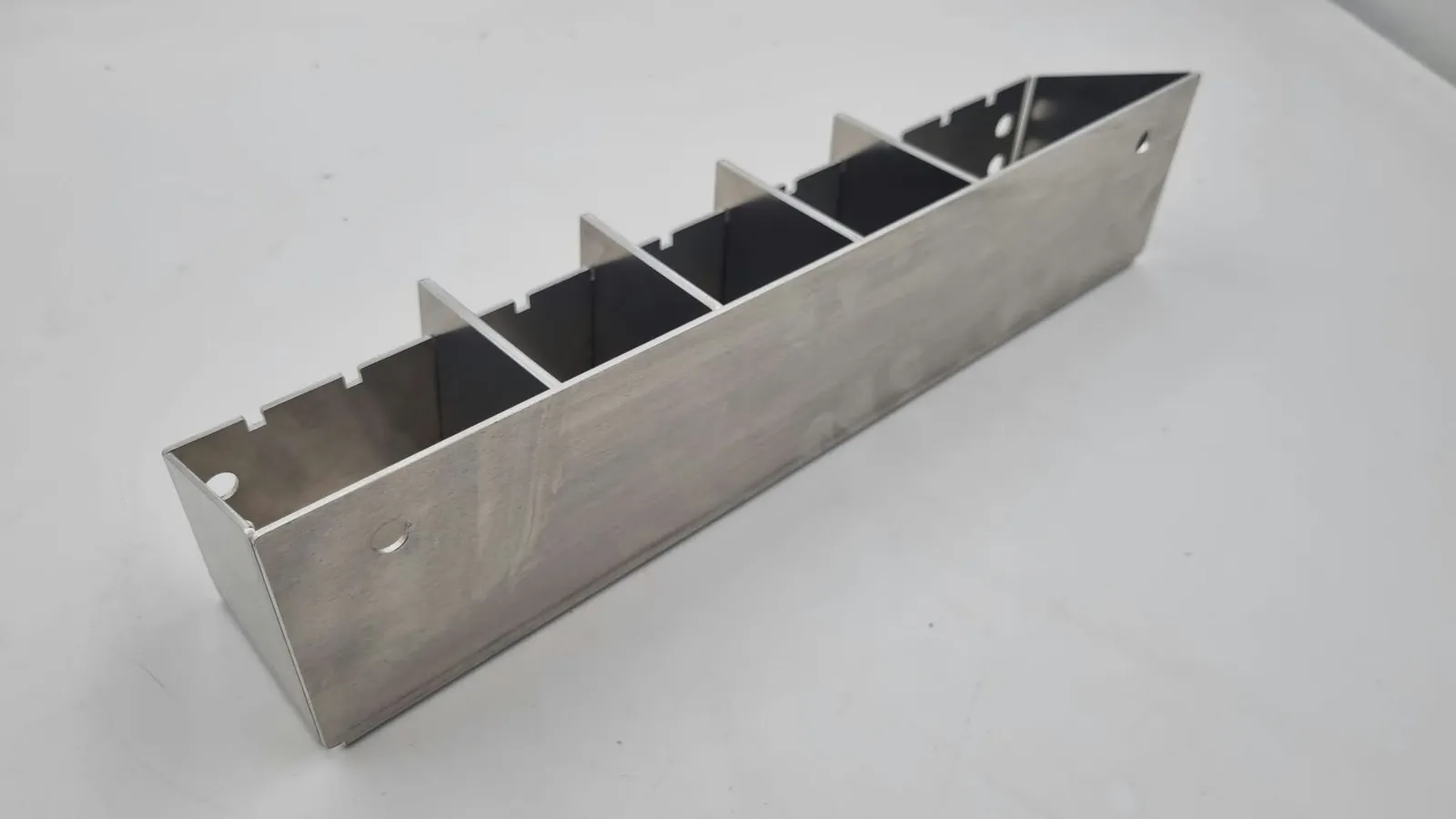 A long open aluminium tray bent from flat sheet, seen from one corner, with four aluminium dividers standing in notches along its sides and round holes near each end