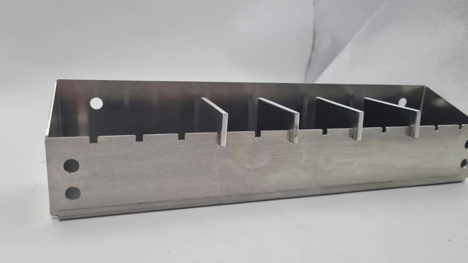 Side view of a bent aluminium tray with evenly spaced notches along its top edge, dividers standing in some of them, and round holes near each end