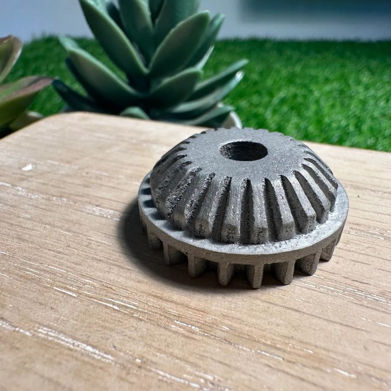 A grey metal bevel gear with a centre hole on a wooden board, straight from the metal 3D printer, its teeth and top face covered in a fine, sandy grain
