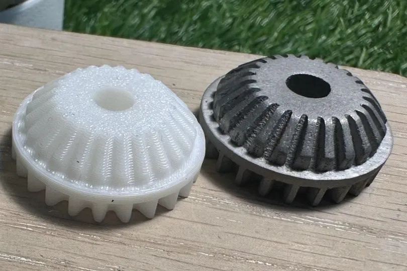 Two copies of the same bevel gear side by side on a wooden board: on the left a white plastic 3D print, on the right the grey metal 3D print with its grainy surface