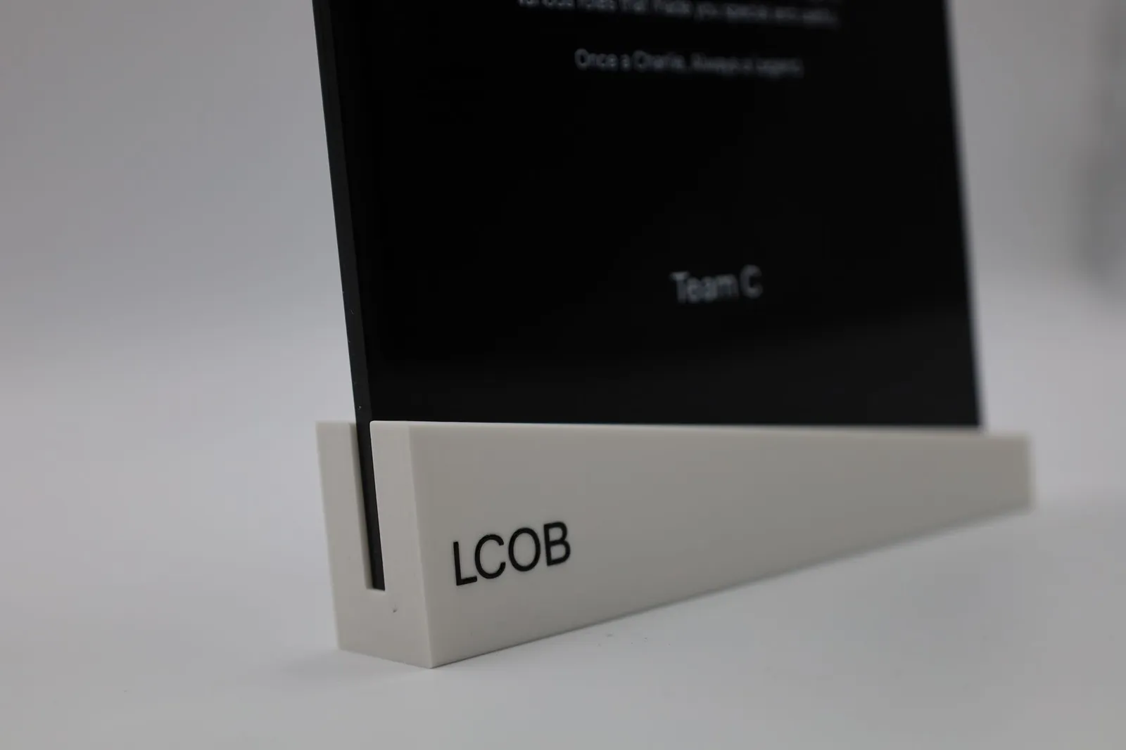 Close view of a black acrylic plaque with small white text printed on it, standing in a white base printed with the letters LCOB in black