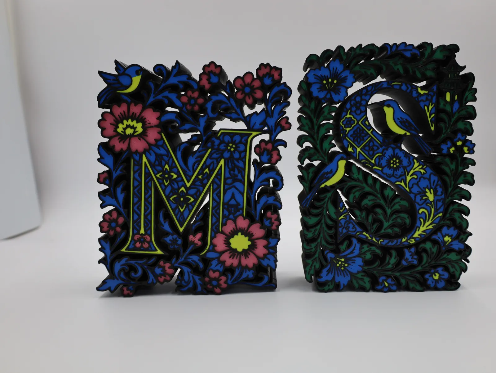 Two ornate letters, M and S, printed in blue, green, pink and lime filament over a black floral relief, standing side by side