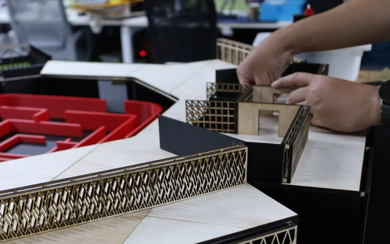 Hands fitting a laser-cut plywood wall with a lattice pattern onto a scale model on a workbench in our studio, with desks and shelves behind