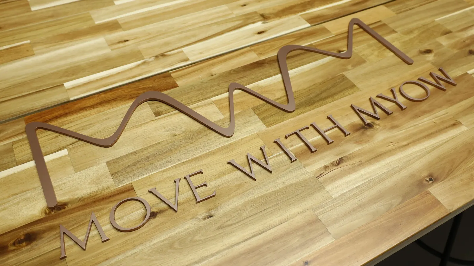 A wavy logo mark and the words Move With Myow in rose gold acrylic letters laid out on a wooden tabletop