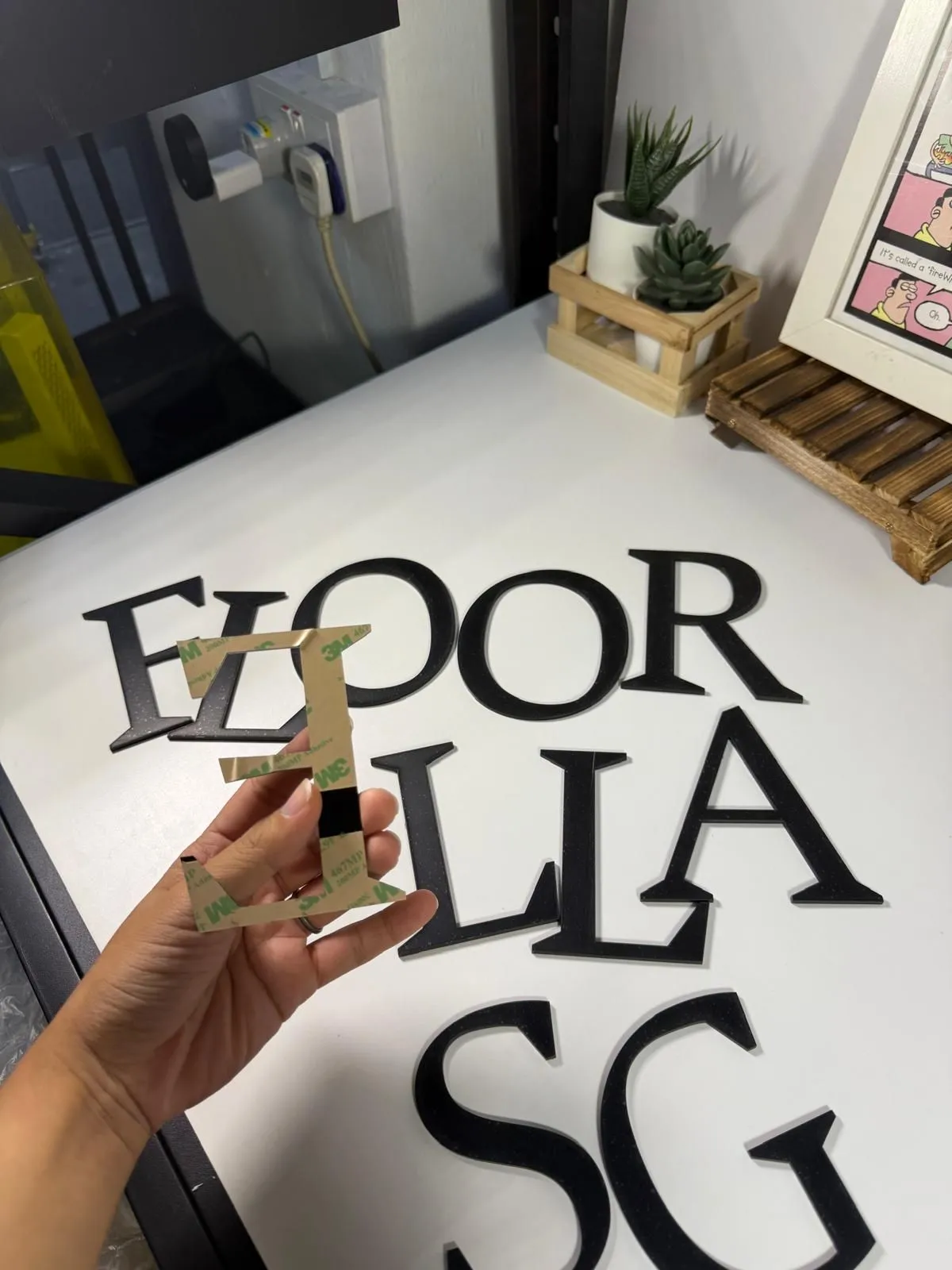 A hand holding a black acrylic letter with 3M adhesive tape on its back, above the other letters of the words Floorella SG laid out on a white table