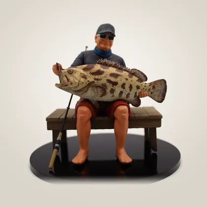 A hand-painted figurine of an angler on a jetty bench, holding up a large spotted grouper