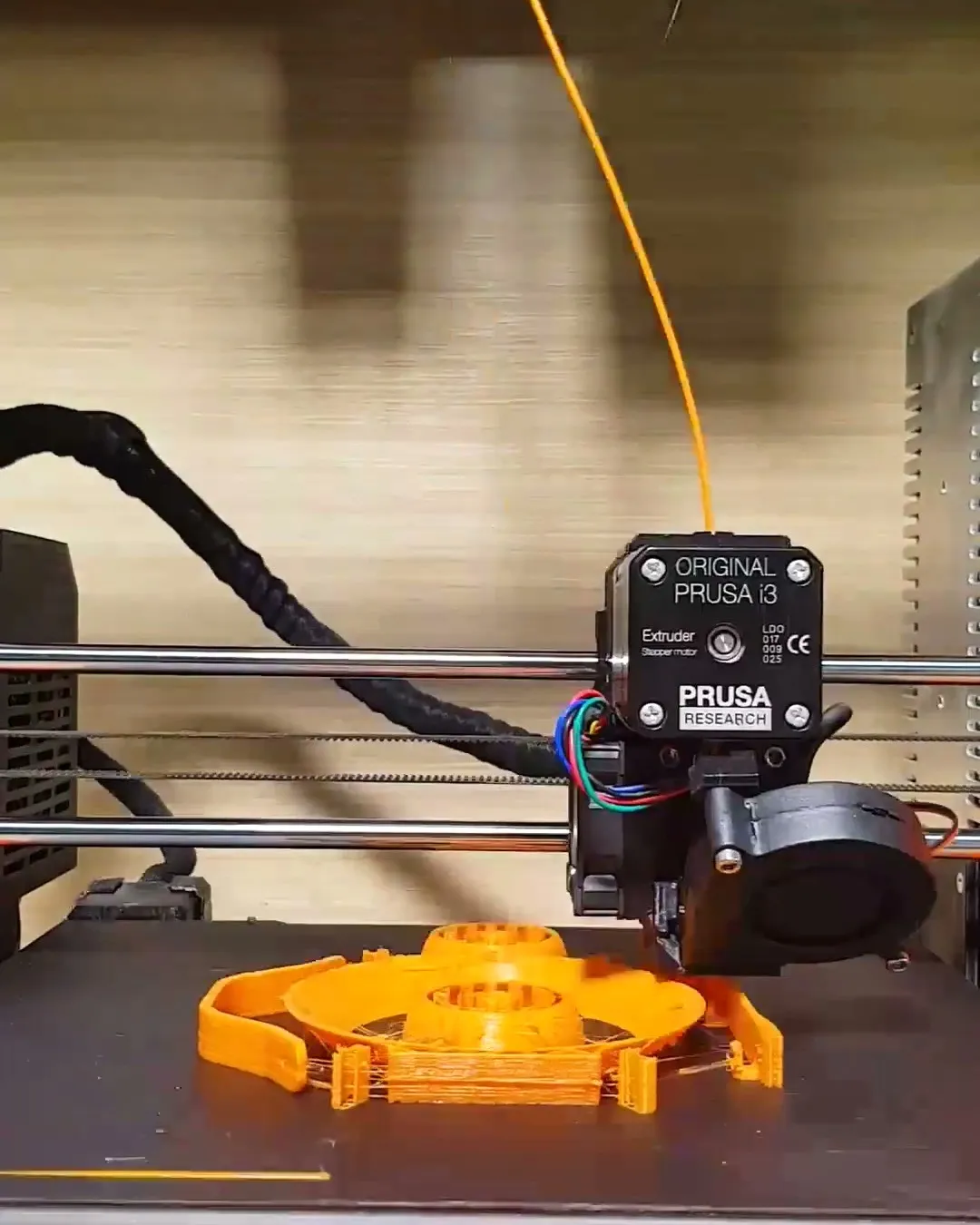 An FDM printer laying down the orange ring and feet of the stand on its bed, with orange filament feeding in from above