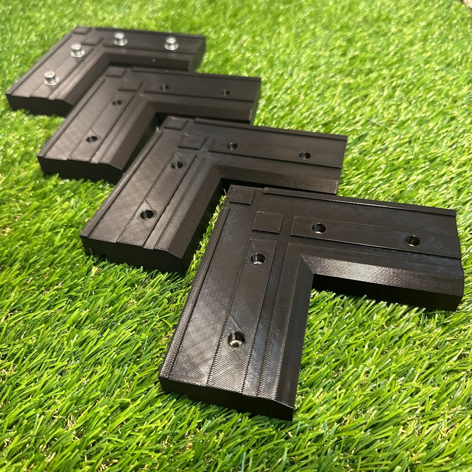 Four black 3D printed replacement L-brackets lined up on artificial grass, the nearest one showing its grooves and fixing holes