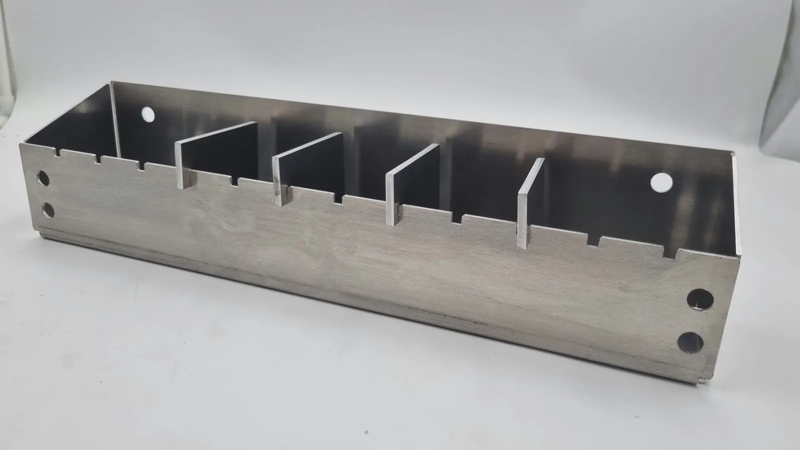 A finished raw aluminium holder from the front: a long folded tray with four flat dividers standing in notches along its front wall, and round holes at each end