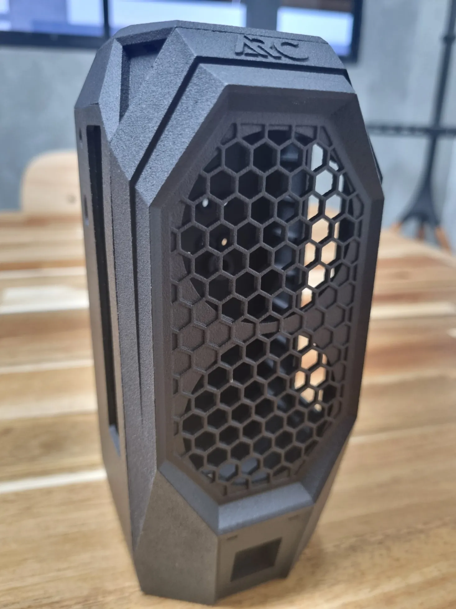 A black nylon computer case standing on a wooden table, with a honeycomb grille across the front and raised lettering on the top cap