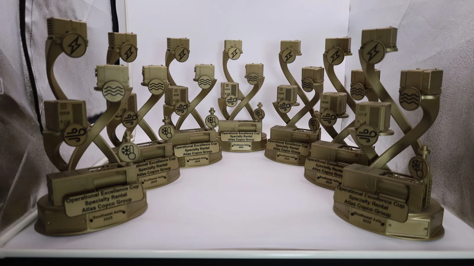 A set of gold Operational Excellence Cup trophies in a light tent, each with miniature machines on two twisting arms and a plate reading Atlas Copco Group