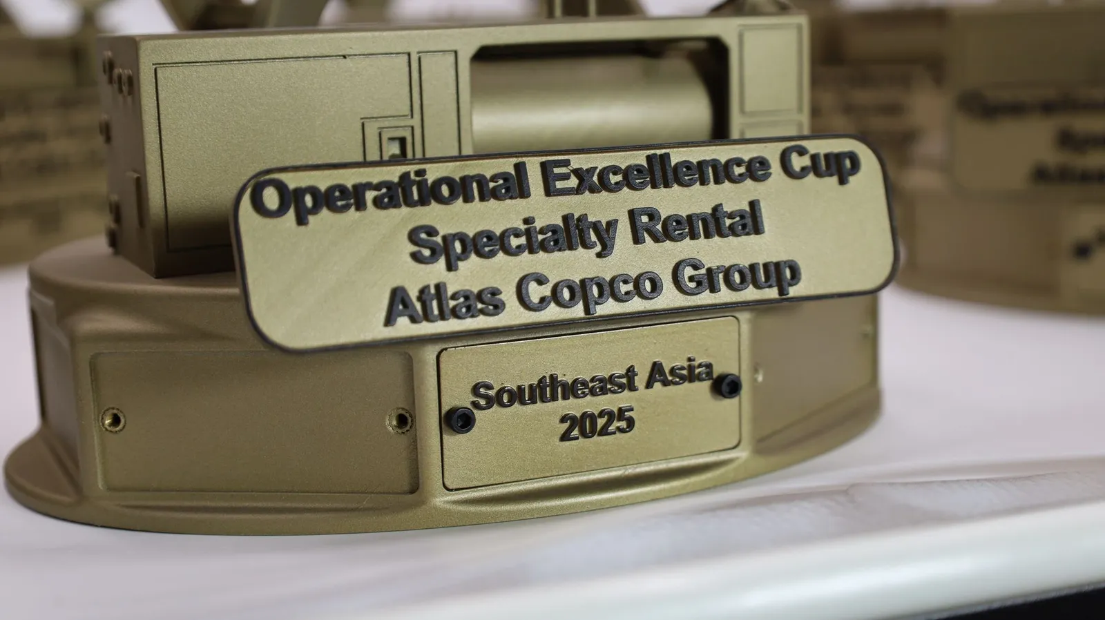 Close-up of the base: a gold plate with raised black lettering reading Operational Excellence Cup, Specialty Rental, Atlas Copco Group, above a screwed-on plate reading Southeast Asia 2025