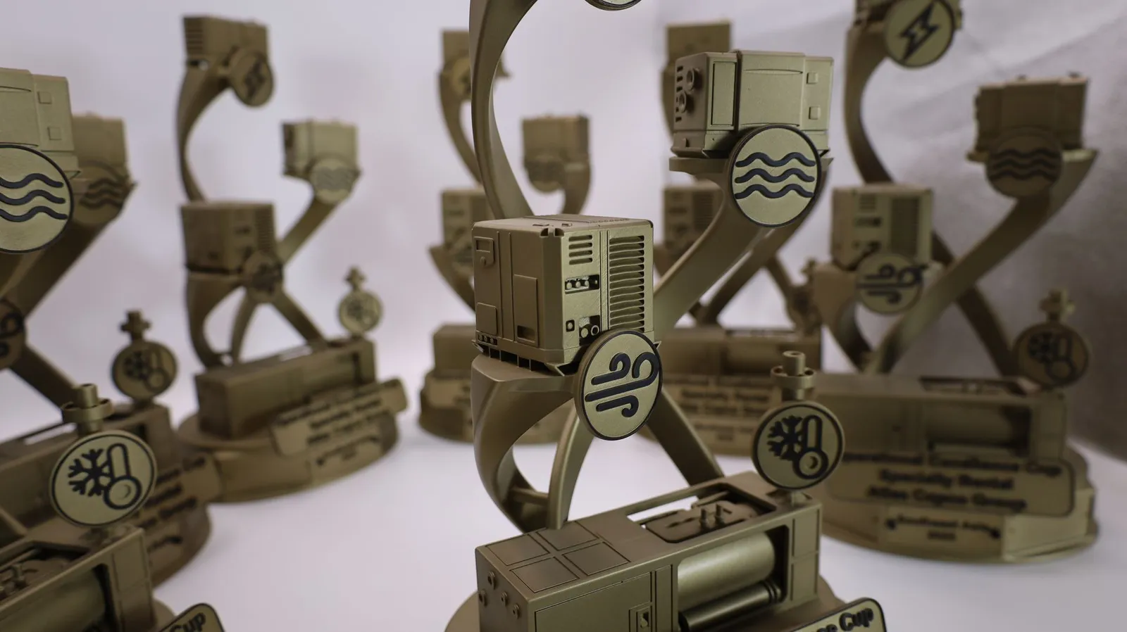 Close-up of the gold trophies: a miniature machine with vents and buttons above a disc with black air lines, and a disc with black waves above