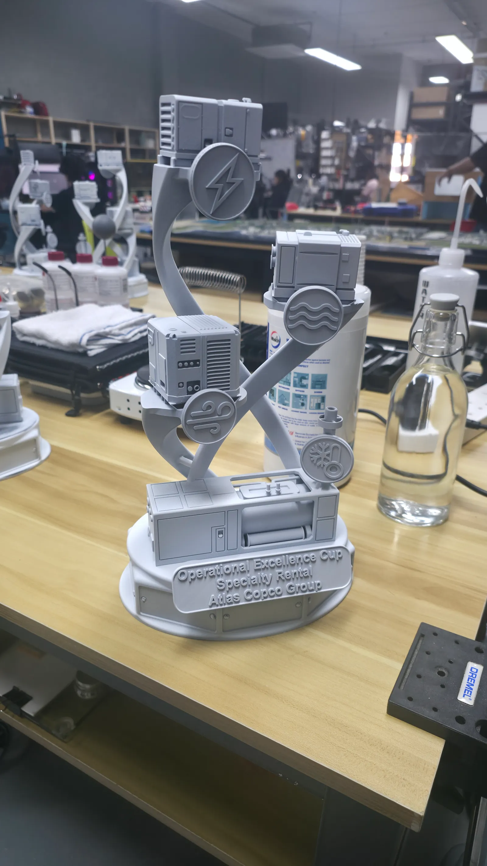 A trophy in plain grey primer on a workbench: miniature machines on two twisting arms, icon discs and a raised plate reading Operational Excellence Cup, Specialty Rental, Atlas Copco Group