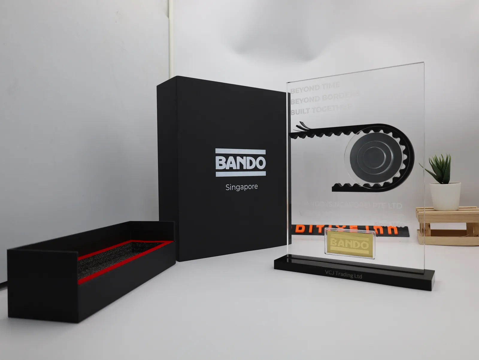 A finished Bando award standing beside its black BANDO Singapore gift box and red-edged foam tray: a black belt wraps a silver pulley on a clear engraved panel above a gold BANDO bar