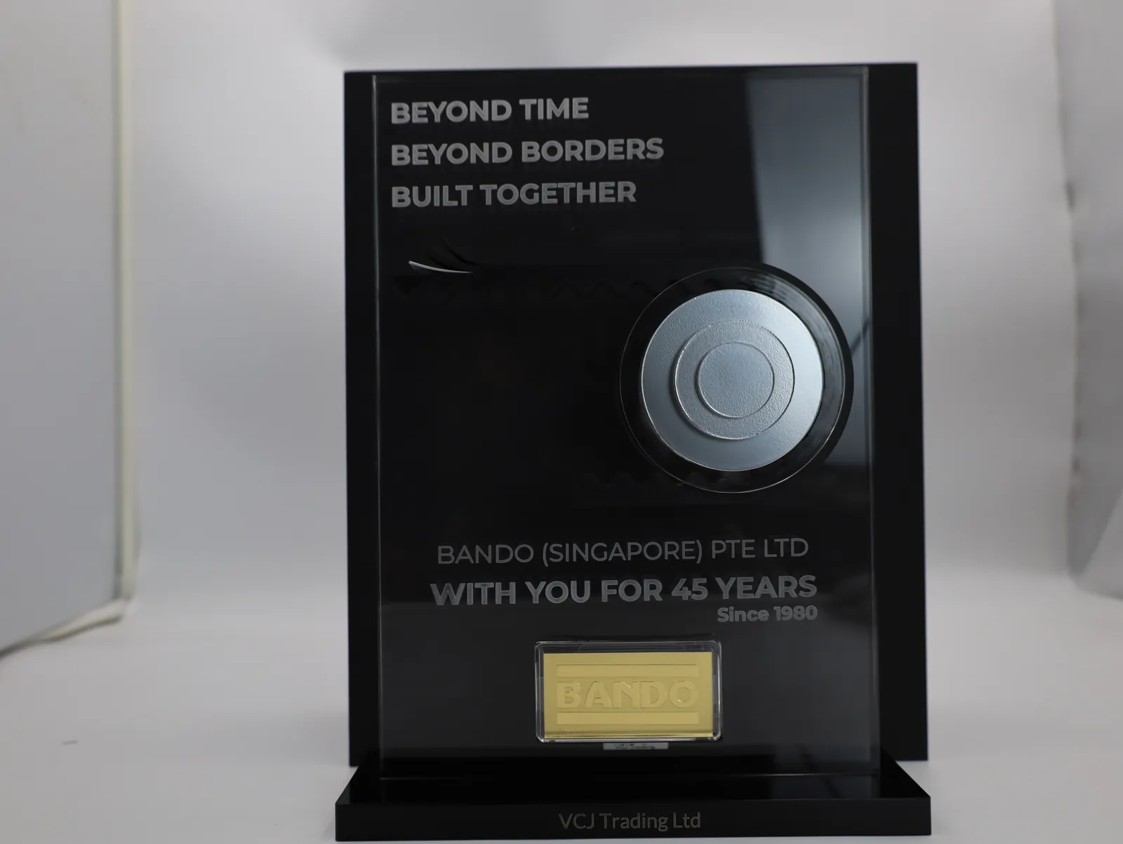 A finished award from the front against its black gift box: engraved wording, the belt round a silver pulley, With you for 45 years, the gold BANDO bar and VCJ Trading Ltd on the base
