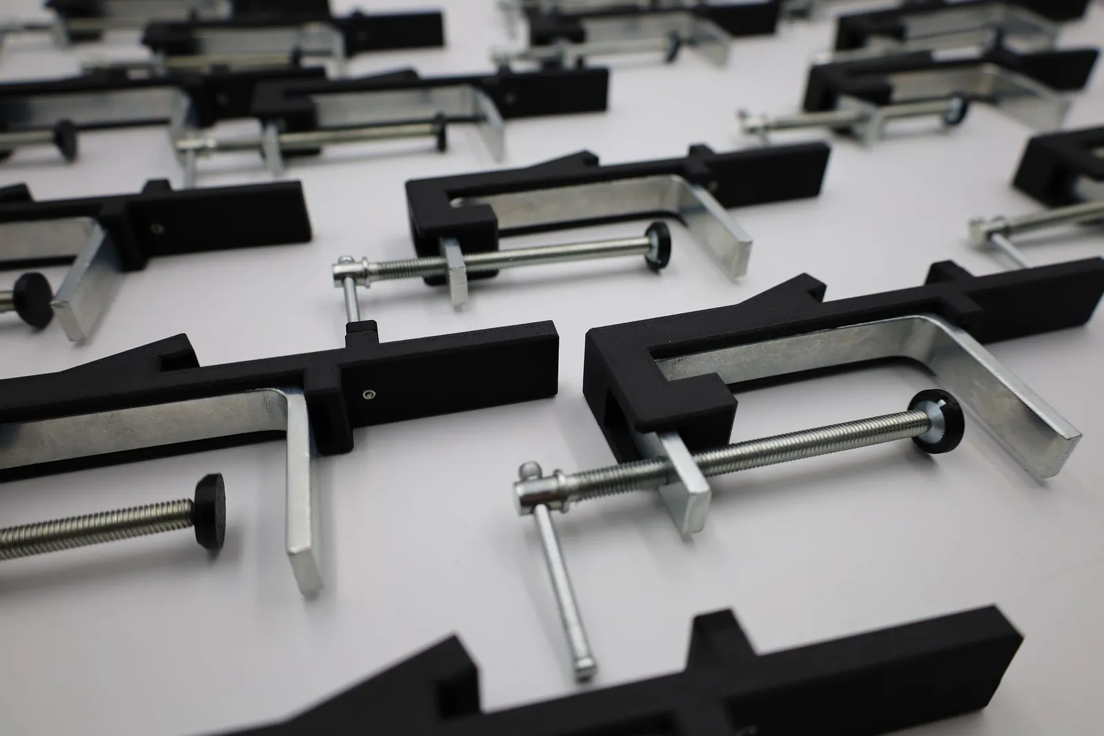 Rows of the finished clamps on a white table: matte black 3D printed arms on galvanised steel screw frames with T-handles