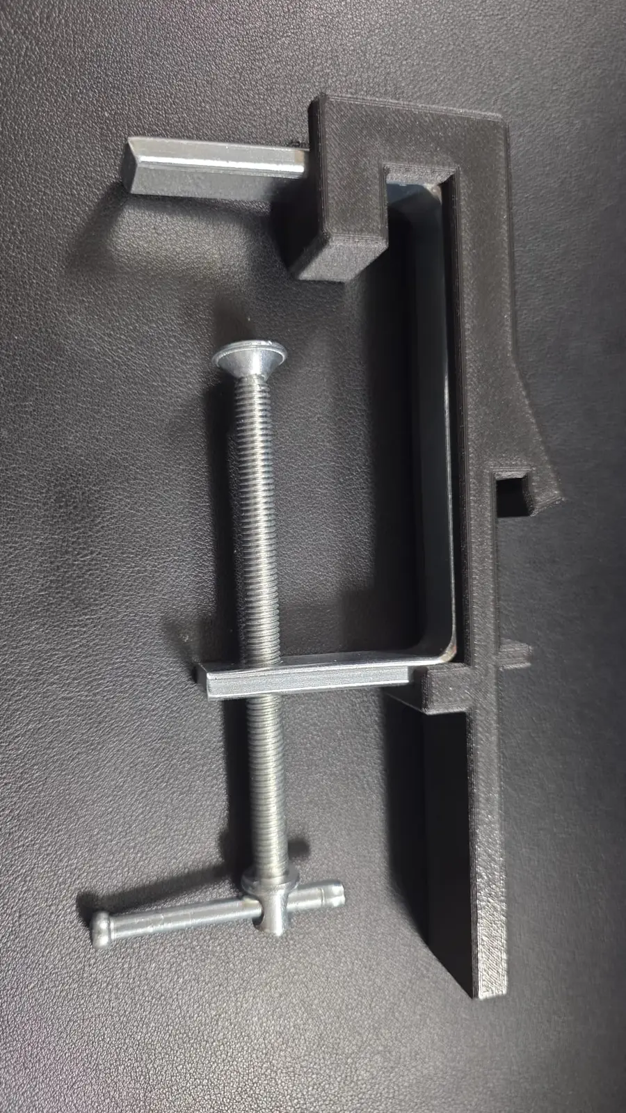 The first printed prototype: a dark grey PETG-CF arm fitted to a steel C-clamp frame with a T-handle screw, on a black mat