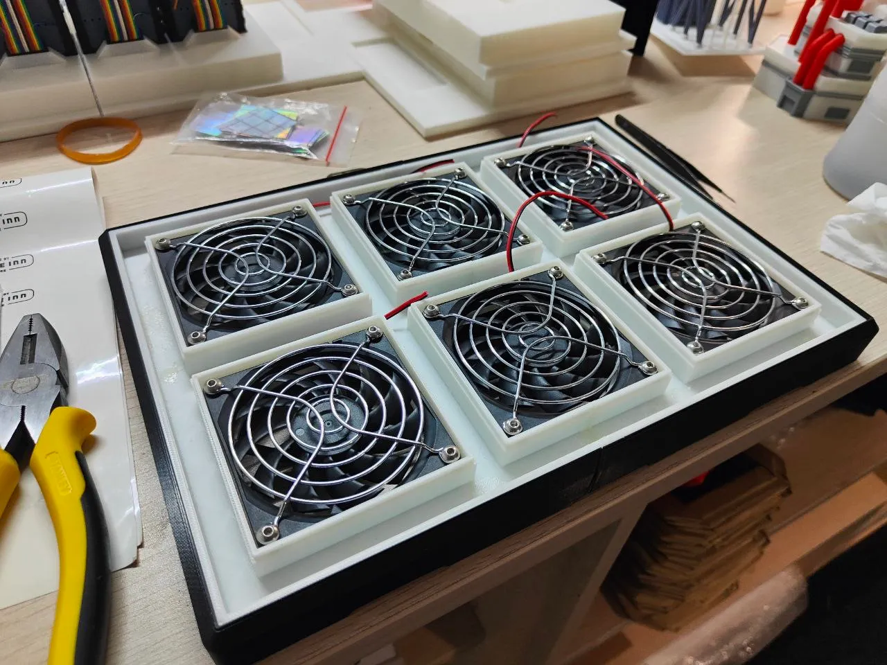 The six fans being fitted into white printed frames on the workbench, wires loose, with printed parts for the model behind
